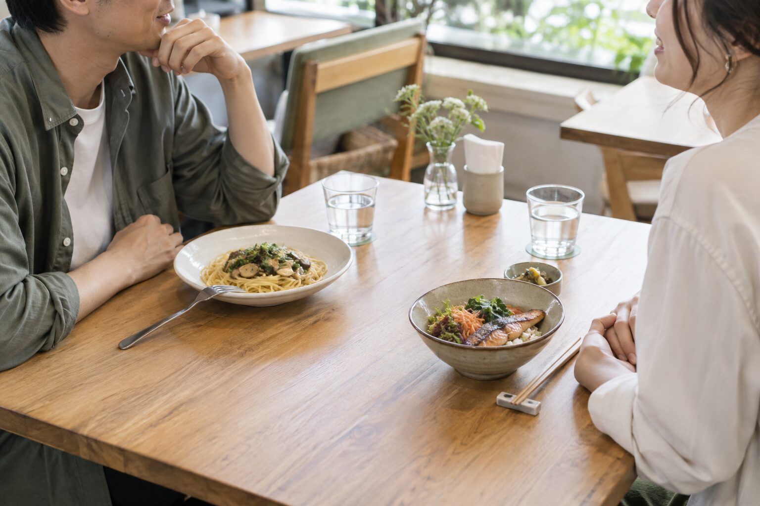 明るいカフェでパスタと魚のご飯をそれぞれ選び、向かい合って話す二人